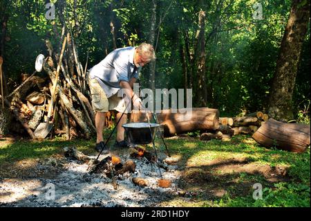 Uomo anziano che prepara cibo sul fuoco da campeggio in pentola smaltata Foto Stock