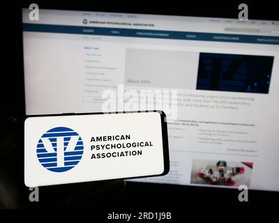 Persona che possiede uno smartphone con il logo dell'American Psychological Association (APA) sullo schermo di fronte al sito Web. Concentrarsi sul display del telefono. Foto Stock