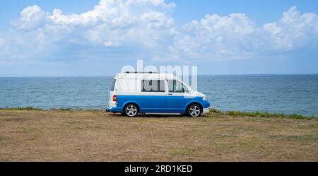 Camper blu vintage parcheggiato su una pianura erbosa con l'Oceano Atlantico sullo sfondo sotto un cielo semi-nuvoloso illuminato dal sole del mattino presto. Foto Stock