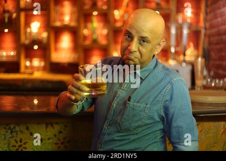 Scena drammatica di un giovane e sfavillante ragazzo calvo in piedi sul bancone del bar. Goditi l'interno del club cittadino mentre il night club regge un bicchiere di whisky Foto Stock