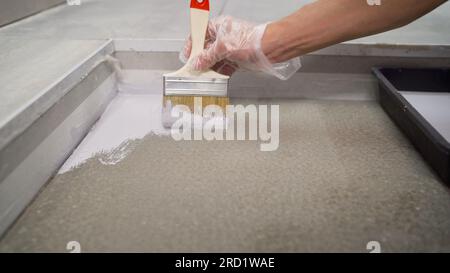 Una mano tiene una spazzola, applicando vernice vernice su una superficie in calcestruzzo. Primo piano, verniciare il pavimento di bianco con una spazzola. Applicazione di un primer bianco sul pavimento Foto Stock