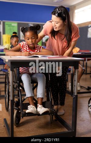 Insegnante femminile felice e diversificata con insegnante di banda in sedia a rotelle che scrive in classe Foto Stock