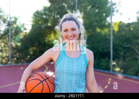 Ritratto di una giocatrice di basket caucasica felice che tiene il basket sul campo da basket Foto Stock