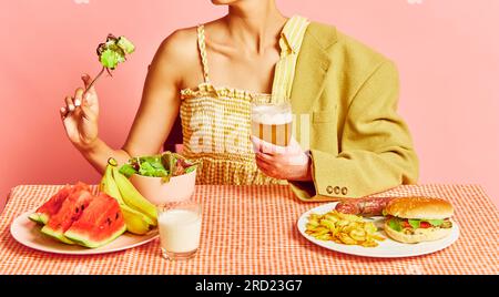 Donna che mangia cibo sano, vitamine, uomo che mangia cibo spazzatura, hamburger e birra sullo sfondo rosa studio Foto Stock