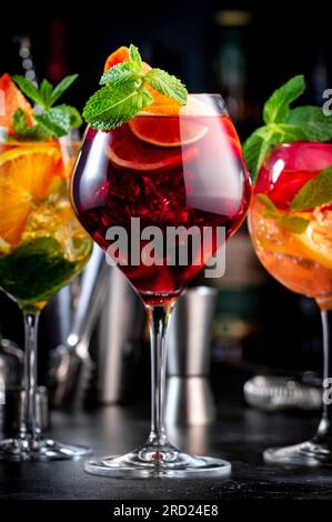 Cocktail alcolici estivi con sangria rosa, rosso, bianco, bevande con vino spagnolo, frutta, agrumi e ghiaccio. Sfondo nero del bancone del bar, utensili da bar in acciaio e. Foto Stock