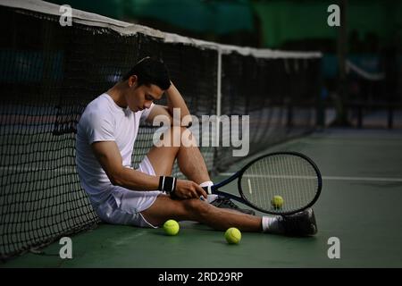 Giocatore asiatico di tennis maschile deluso seduto sul campo, turbato per la perdita di concorrenza, emozioni di fallimento Foto Stock