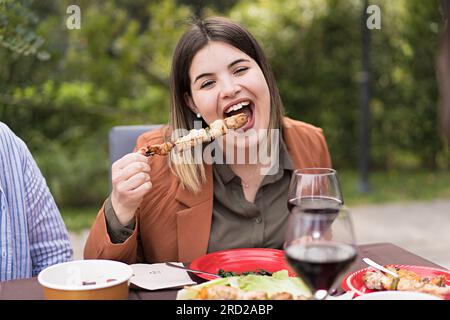 Una donna gioiosa e di grandi dimensioni assaggia uno spiedino barbecue durante un pasto nel tardo pomeriggio in una villa a Trecastagni. I bicchieri da vino rosso suggeriscono un'atmosfera rilassata e festosa Foto Stock