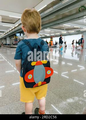 In un aeroporto affollato, un bambino perduto si trova da solo, con uno zaino, guardando fuori dalla finestra gli aeroplani. La triste espressione del ragazzo suggerisce la sua stanchezza e isolamento tra la folla affollata. Foto Stock