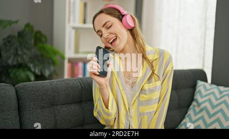 Giovane donna bionda che ascolta la musica seduta sul divano cantando canzone a casa Foto Stock