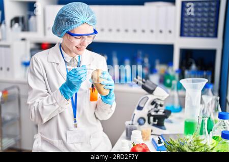 Giovane scienziata bionda che tiene in laboratorio un campione di patata con pinzette Foto Stock