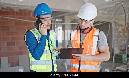 Due uomini costruttori scrivono un documento che parla su smartphone in cantiere Foto Stock