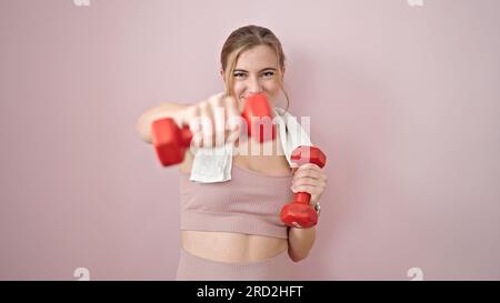 Giovane donna bionda che indossa abbigliamento sportivo da boxe con manubri su sfondo rosa isolato Foto Stock