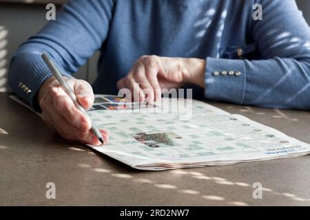 Primo piano delle mani di una donna anziana, seduta a un tavolo in casa sua, risolvendo un puzzle di parole incrociate. Terapia di riabilitazione cognitiva per anziani. selettivo foc Foto Stock