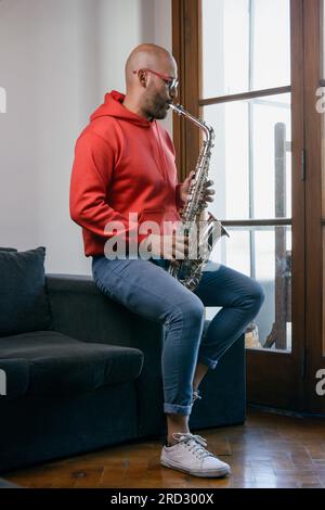 immagine verticale di un giovane sassofonista latino vestito di rosso con barba e bicchieri, che gioca a casa seduto sul divano del soggiorno Foto Stock
