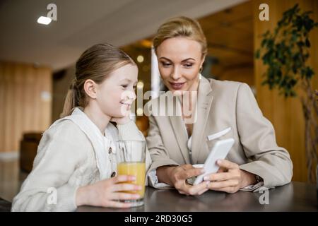 madre e figlia felici che trascorrono del tempo insieme, donna bionda che tiene lo smartphone vicino a una figlia allegra, era digitale, genitore che lavora e chil Foto Stock