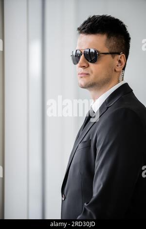 copricapo professionale, protezione del corpo con auricolare, bell'uomo con occhiali da sole e tuta nera con cravatta, sicurezza in hotel, gestione della sicurezza, topografo Foto Stock