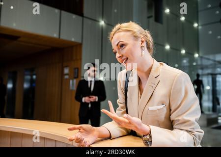 donna bionda educata con i gesti mentre parla alla reception, concetto di servizio di sicurezza personale, guardia del corpo in tuta in piedi sul retro sfocato Foto Stock