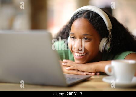 Donna nera felice che indossa le cuffie per guardare contenuti multimediali sul computer portatile in una terrazza del bar Foto Stock