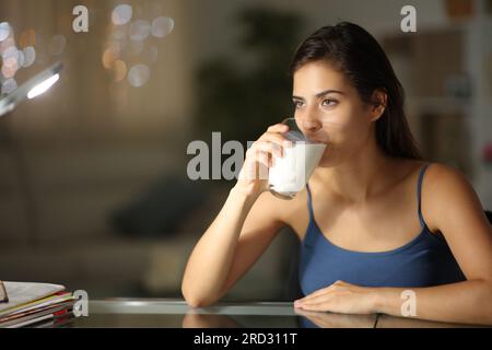 Donna di notte che beve latte a casa guardando altrove Foto Stock