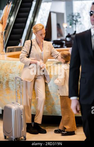 concetto di sicurezza personale, donna bionda in piedi alla reception e parla con la figlia preadolescente, stile di vita ricco, viaggi in famiglia, guardia del corpo in tuta a. Foto Stock