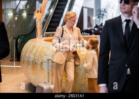 concetto di sicurezza personale, donna bionda in piedi al banco della reception con una figlia preadolescente, stile di vita ricco, viaggi in famiglia, guardia del corpo in costume e vetro solare Foto Stock