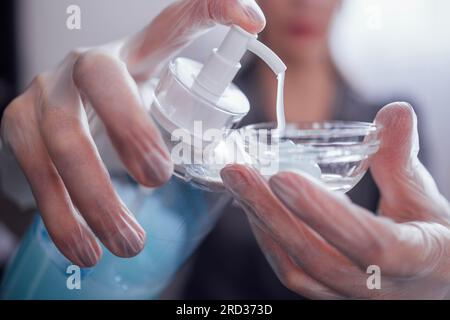 Primo piano delle mani femminili in guanti trasparenti che strizzano il gel blu in una ciotola di vetro. Il cosmetologo si sta preparando per una procedura cosmetica e i quadranti Foto Stock