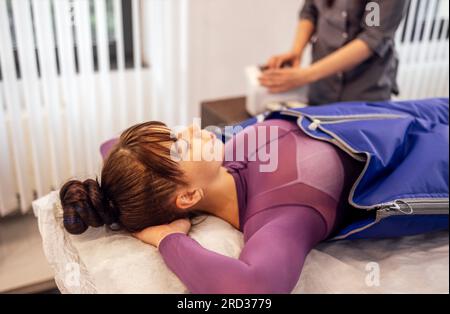 Una giovane donna attraente giace con le mani dietro la testa in un vestito speciale durante la pressoterapia. Massaggio di drenaggio linfatico hardware in un salone termale Foto Stock