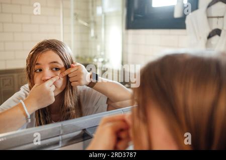 Una ragazza adolescente carina tira fuori un brufolo sulla guancia in bagno. Un'adolescente in abiti casual con una pelle problematica tocca il suo viso. Riflessione in t Foto Stock