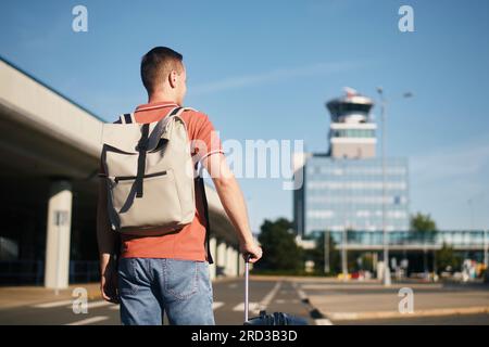 Viaggio in aereo. Il viaggiatore è a pochi passi dal terminal dell'aeroporto. Uomo con valigia contro la torre di controllo del traffico aereo. Foto Stock