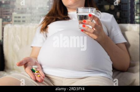 Una ragazza incinta tiene una tazza d'acqua e una manciata di pillole nel palmo. Antidolorifici e vitamine durante la gravidanza. Primo piano Foto Stock