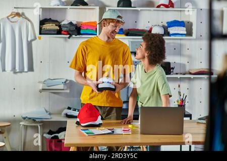Giovani designer multietnici positivi che parlano mentre lavorano con istantanee e notebook vicino a campioni di tessuti sul tavolo in uno studio di stampa, un'azienda sostenibile Foto Stock