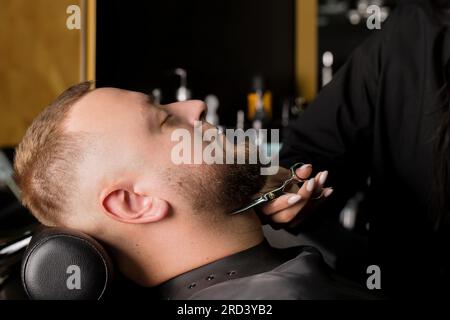 La bella giovane donna, il grande parrucchiere maschile, taglia la barba con le forbici nel barbiere Foto Stock