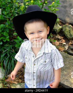 Bambino, che indossa un cappello da cowboy nero in feltro, regala un radioso, sorriso in trasmissione. Ha una stampa bianca, una camicia da cowboy anteriore. Foto Stock