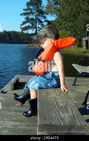 Un bambino è seduto su un molo di legno e indossa un giubbotto di sicurezza per l'acqua. Indossa galosci di gomma e tute. La sua lingua è tra le labbra come se fosse tastin Foto Stock