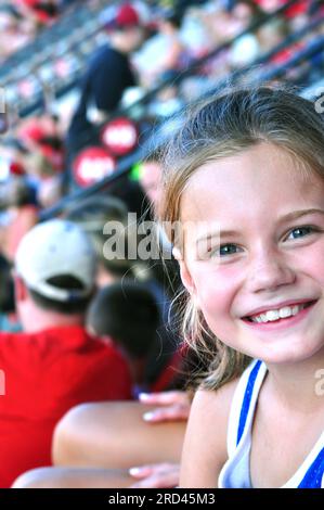 La bambina sorride la sua eccitazione mentre si siede al suo primo gioco professionale di baseball. Si siede negli stand con la sua famiglia. Foto Stock