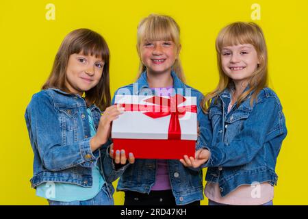 Ragazze adolescenti che presentano una confezione regalo di compleanno con nastro rosso che si allunga le mani offrono un regalo avvolto che celebra la festa e sconti pubblicitari per le festività. Bambine sorelle. Tre fratelli e figli Foto Stock