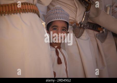 Un giovane ragazzo vestito con un tradizionale dishdasha dell'Oman e un cappello Zanzibari cova una folla di uomini adulti mentre si esibiscono in una danza. Foto Stock