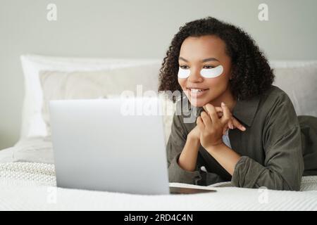 Una ragazza di razza mista si rilassa con cerotti oculari, orologi serie su laptop. Pubblicità per la cura della pelle. Foto Stock