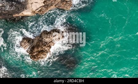 Vista aerea direttamente sopra le onde bianche che si infrangono su aspre rocce di pietra in un ambiente aspro sulla costa della Cornovaglia con spazio copia sopra il verde smeraldo Foto Stock