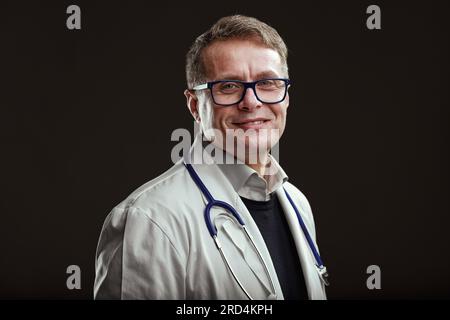 Un medico di mezza età sorride con gli occhi, appassionato della sua professione, gentile e contento di aiutare le persone attraverso la sua conoscenza. Sfondo scuro Foto Stock