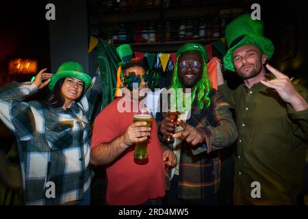 Gruppo di giovani amici interculturali in abito St Patrick che guardano la macchina fotografica mentre sono in fila, tifo con un bicchiere di birra e si divertono con la festa Foto Stock