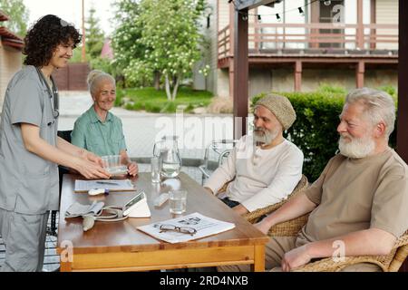 Felice giovane infermiera che porta medicinali a un gruppo di anziani seduti a tavolo sulla terrazza della casa di riposo la mattina d'estate Foto Stock