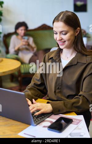 Giovane donna d'affari moderna con abbigliamento casual intelligente che digita sulla tastiera del computer portatile mentre si siede a un tavolo in un bar contro una collega Foto Stock