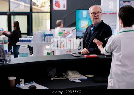 Farmacia cliente anziano che guarda la cassiera che effettua la scansione dell'acquisto presso la reception, acquista crema solare e supplemento cardiaco in farmacia. Uomo anziano con occhiali in piedi alla scrivania e in attesa di pagare Foto Stock