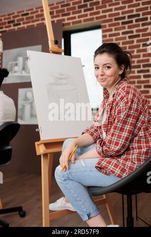 Una giovane donna felice sorridente alla macchina fotografica mentre è seduta a cavalletto durante una divertente lezione di arte creativa, imparando le basi del disegno e provando qualcosa di nuovo. Studentessa che si diverte ad attività artistiche Foto Stock