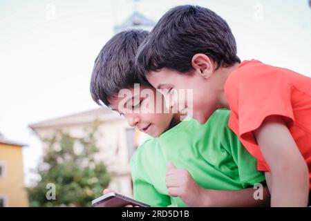 due ragazzi che si divertono a mandare messaggi con un telefono cellulare, due ragazzi con magliette verdi e rosse che ridono con uno smartphone in mano Foto Stock