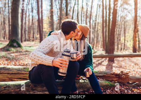 Coppia felice che si bacia bevendo tè dal thermos e si rilassa nella foresta autunnale. Uomo e donna seduti sul tronco che bevono un caffè dalla beuta sottovuoto. Peopl Foto Stock