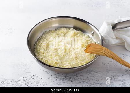 Padella con riso arborio, cipolla e brodo su sfondo azzurro. Cucinare risotto fatto in casa con pera e gorgonzola, passo dopo passo, passo 4. Foto Stock