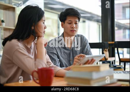 Uno studente universitario asiatico intelligente e concentrato che tiene lezioni e aiuta il suo amico con la matematica mentre studia in uno spazio di co-working in un caffè. Foto Stock