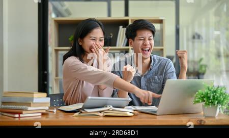Due giovani studenti universitari asiatici allegri e felici guardano lo schermo di un notebook, ridono e celebrano insieme il successo del loro progetto scolastico Foto Stock
